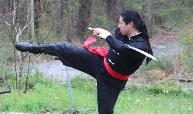 John in a Chinese long shirt holding a broadsword and executing a kick. 